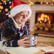 Junge liegt mit einer Tasse Kakao auf dem Teppich und liest ein Buch, der Hintergrund ist weihnachtlich
