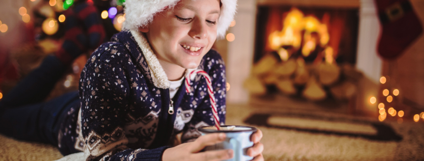 Junge liegt mit einer Tasse Kakao auf dem Teppich und liest ein Buch, der Hintergrund ist weihnachtlich